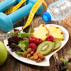 A bottle of water, dumbbells and a measuring tape are next to a plate of fruit and veg.