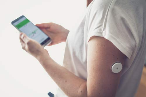 Side view of the upper body of a woman. A CGM sensor is secured to her left upper arm and she is holding a mobile phone in her hands, with a blurred glucose chart.