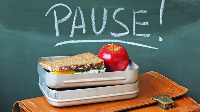 Auf einer Aktentasche steht eine offene Brotdose mit einem Vollkornbrot und einem Apfel. Im Hintergrund steht an einer Tafel das Wort "Pause".