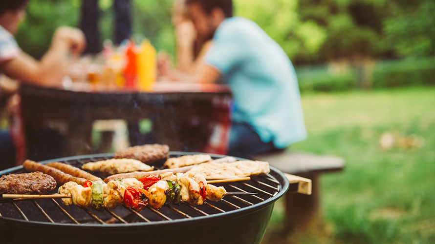 Der neue Diabetes-Spickzettel für das Grillen zeigt die Kohlenhydratmengen beliebter Grillspeisen, Beilagen, Saucen und Getränke auf einen Blick. © bernardbodo / Adobe Stock Im Vordergrund ist ein Grillrost mit Gemüsespießen und Fleisch zu sehen, im Hintergrund sitzen Menschen an einem Tisch im Park. Auf dem Tisch stehen Saucen.