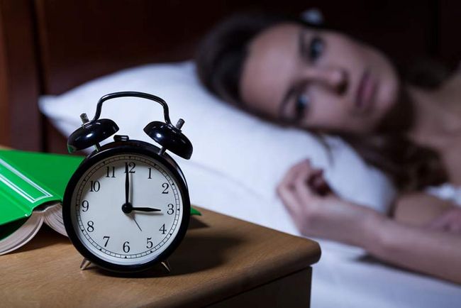 Eine Frau liegt wach in ihrem Bett. Im Vordergrund steht ein Wecker, der 3 Uhr anzeigt. 