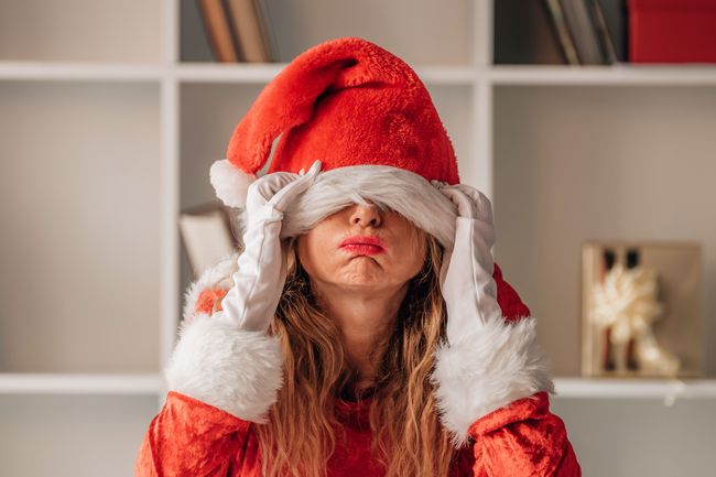 Eine Frau sitzt in einem weihncahtlichen Kostüm am Tisch und ziehst sich gestresst die Weihnachtsmütze über die Augen. 