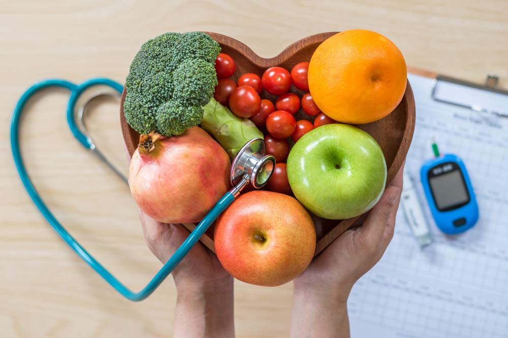 Zei Hände halten eine herzförmige Schale mit Obst und Gemüse, im Hintergrund sieht man ein Blutzuckermessgerät und ein Stetoskop.