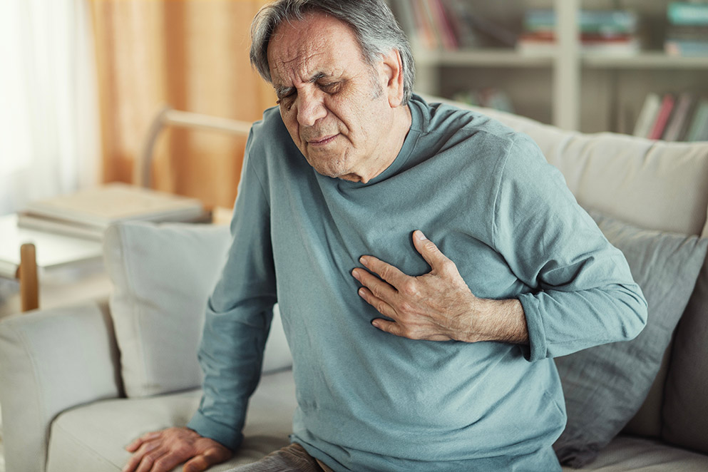 Ein älterer Mann sitzt auf dem Sofa und greift sich mit der linken Hand ans Herz.